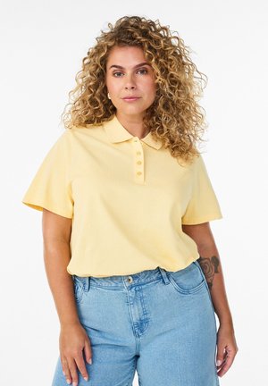 Žena s kučeravými vlasmi, oblečená v svetložltom krátkoročnom polotričku a modrých džínsach s vysokým pásom, stojaca s neutrálnym výrazom.