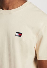 Beige košile s krátkým rukávem a malou obdélníkovou nášivkou s logem "Tommy Jeans" v námořnické modré, bílé a červené barvě na hrudi.