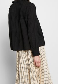 Svart långärmad topp med vertikala veck och hög krage, parat med en beige kjol med svart rutig mönster.