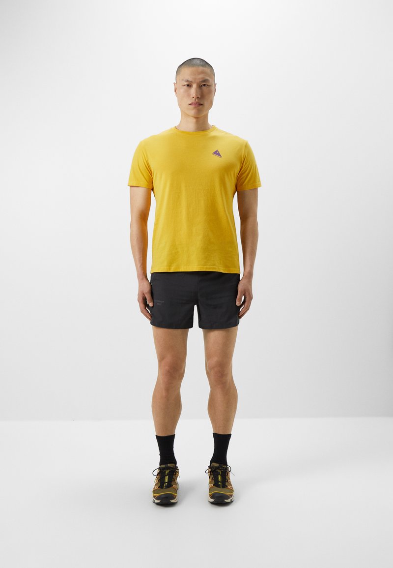 Maglietta di cotone gialla con un piccolo logo viola, abbinata a pantaloni corti neri e sneakers gialle e nere, su uno sfondo neutro.