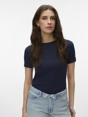 Mladá žena s dlhými hnedými vlasmi, oblečená v tmavomodrom tričku s krátkym rukávom a svetlomodrých džínsach s vysokým pásom na jednoliatom pozadí.