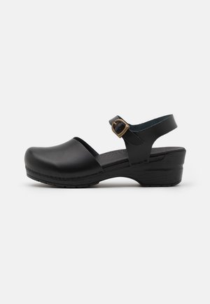 Black clogs with a rounded toe and an ankle strap featuring a bronze buckle. Made of smooth leather with a low, textured wedge sole.