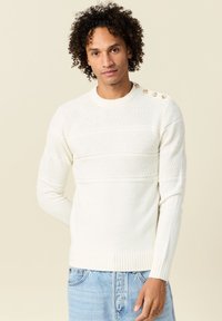 Maglione bianco a maglia con strisce testurizzate, colletto rotondo e dettagli in bottone sulla spalla, abbinato a jeans azzurri chiari.