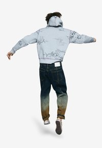 Szürke kapucnis pulóver nyomtatott betűs mintákkal, kék farmerrel párosítva, amely sötéttől világosig terjedő színátmenetet mutat. Alatta sportcipők láthatók.