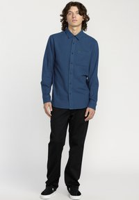 Chemise bleue à manches longues avec un col boutonné, une poche poitrine et des boutons noirs, assortie à un pantalon noir et des chaussures noires à enfiler.