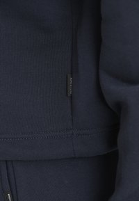 Marineblauwe katoenen blend kledingstuk met een gladde, gestructureerde oppervlakte. Kenmerkt zich door een zwart label met "ENDURANCE" op de naad. Close-up van de stikdetails.