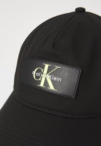 Zwarte baseballcap van textuurstof met een prominent rechthoekig logo-embleem waarop de merknaam in groen en wit staat.