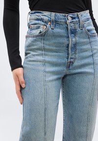 Γυναίκα που φοράει ψηλόμεσα ανοιχτό μπλε τζιν με κάθετες ραφές και μαύ ρο μακρυμάνικο τοπ, με το χέρι ακουμπισμένο στο μηρό.