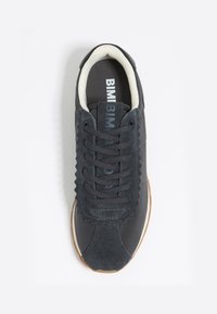 Zapatilla gris oscuro con materiales de ante y malla, presentando un borde en forma de ola y cordones redondos. Suela de goma color tostado con logo en la lengüeta.