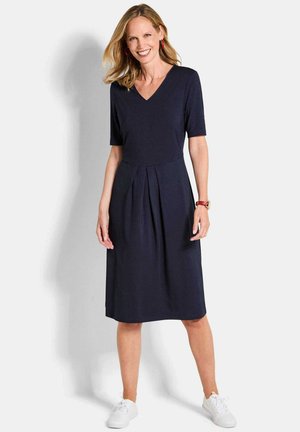 Femme souriante, portant une robe bleu marine plissée jusqu'aux genoux, des baskets blanches, des boucles d'oreilles rouges en créole et une montre-bracelet rouge, posant devant un fond blanc.