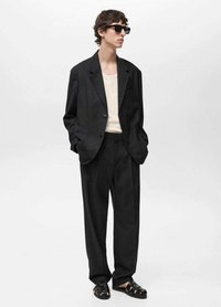 Chaqueta de traje negra oversized con pantalones de pierna ancha, combinada con una blusa beige y sandalias negras. Presenta un solo botón y detalles en los bolsillos.