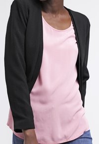 Cardigan nero aperto davanti indossato su una blusa rosa con una trama morbida. Il cardigan ha maniche lunghe e una vestibilità rilassata, adatta per sovrapposizioni.