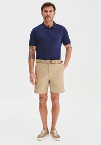 Granatowa koszulka polo, beżowe szorty, brązowy pasek, casualowe sneakersy. Koszulka polo ma kołnierzyk i krótkie rękawy, a szorty sięgają do kolan.