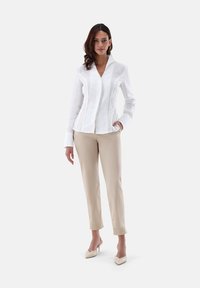 Camicia bianca in lino con colletto, abbinata a pantaloni beige e scarpe con tacco crema. Tessuto liscio, design aderente.