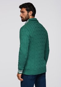 Suéter verde de punto con un alto cuello y puños a rayas, con una superficie texturizada y un dobladillo acanalado, usado sobre jeans oscuros.