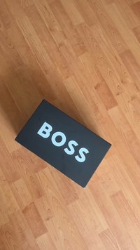 Czarne prostokątne pudełko z matowym wykończeniem, na górze wyraźnie widoczne białe słowo "BOSS". Pod spodem widoczna drewniana podłoga.