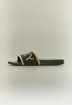 Sandale mule unique vert foncé avec bord beige et lettres blanches partielles sur la bride texturée, montrée de profil sur un fond neutre.