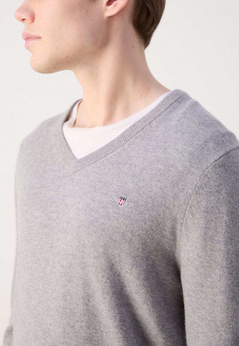 Grau V-Ausschnitt Pullover aus weichem Stoff mit subtiler Textur; verfügt über ein kleines gesticktes Logo auf der Brust.