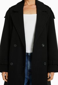 Czarny wełniany płaszcz z dużymi klapami, podwójnym zapięciem i guzikami. Charakteryzuje się dopasowanym krojem i akcentami przy nadgarstkach. Pod spodem biała bluzka.