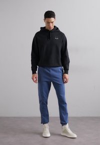Zwarte hoodie met een klein smiley-logo, gecombineerd met blauwe tapering broek en witte sneakers. Soepele texturen, casual pasvorm.