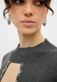 Szary dziany sweter z wysokim kołnierzem z ozdobnymi elementami wzdłuż przedniego dekoltu; modelka nosi srebrne kolczyki w kształcie kół.