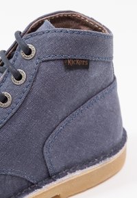 Bottine à cheville en toile bleue avec une étiquette en cuir marron, des œillets en argent et une semelle en caoutchouc. Caractéristiques : tissu texturé et finitions cousues.