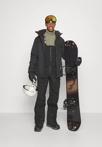 Snowboardåkare i svart jacka och byxor, håller en snowboard och hjälm. Har böjd design, goggles, handskar och fastsatta bindningar.