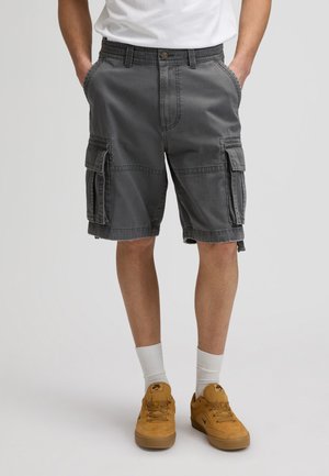 Egy férfi szénszürke terep rövidnadrágot, fehér zoknit és mustár sárga sportcipőt visel, zsebre tett kézzel áll.