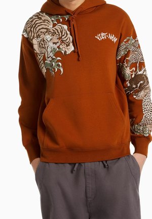 Bluza z kapturem w kolorze rdzy z detalicznymi wzorami tygrysa i smoka na rękawach i na klatce piersiowej, w połączeniu z szarymi spodniami, ręce w kieszeniach.