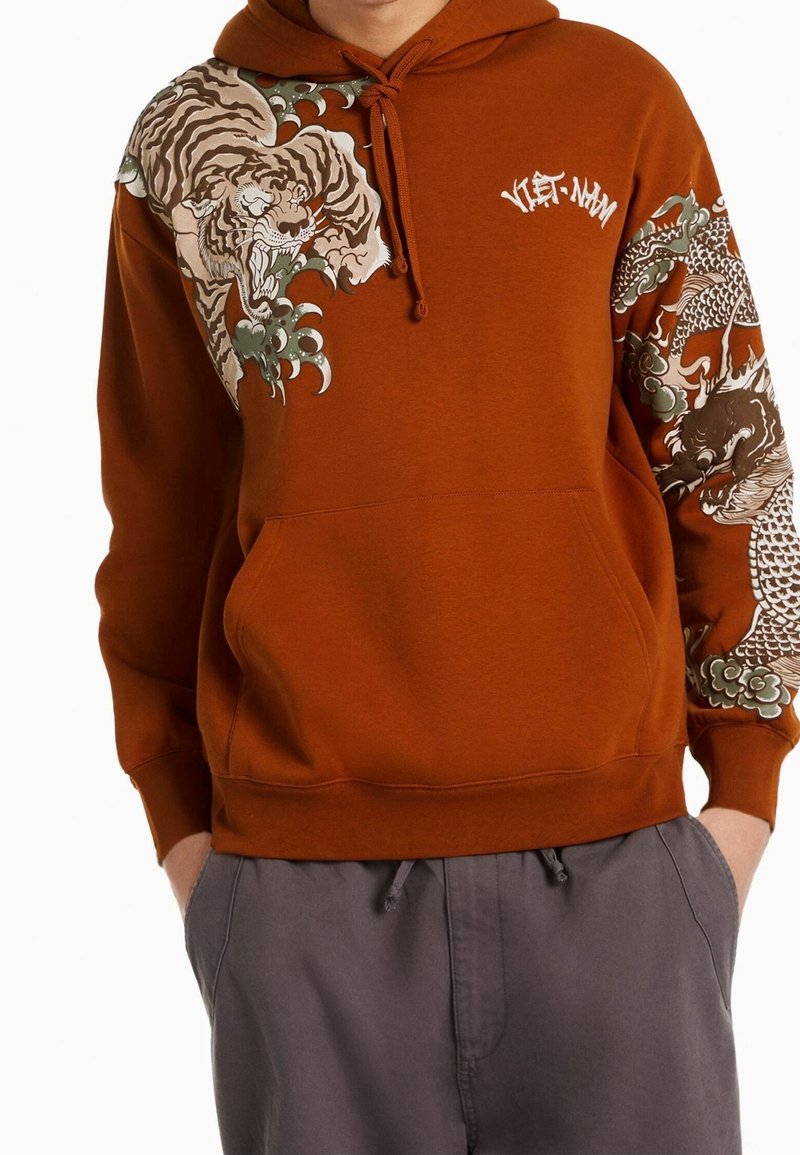 Sweat à capuche couleur rouille avec des motifs détaillés de tigre et de dragon sur les manches et la poitrine, associé à un pantalon gris, mains dans les poches.