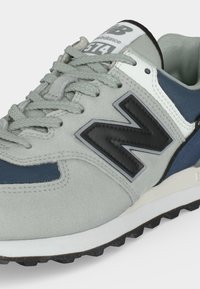 Сиви и сини маратонки New Balance 574 с черно лого, подплънка на яката и текстурирани връзки на бял фон.