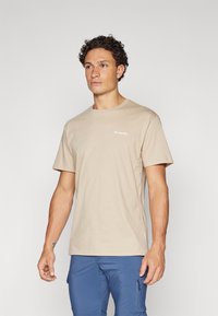 Tricou din bumbac bej, cu mâneci scurte, decolteu rotund și un mic logo Columbia pe partea stângă a pieptului. Purtați cu pantaloni albaștri.