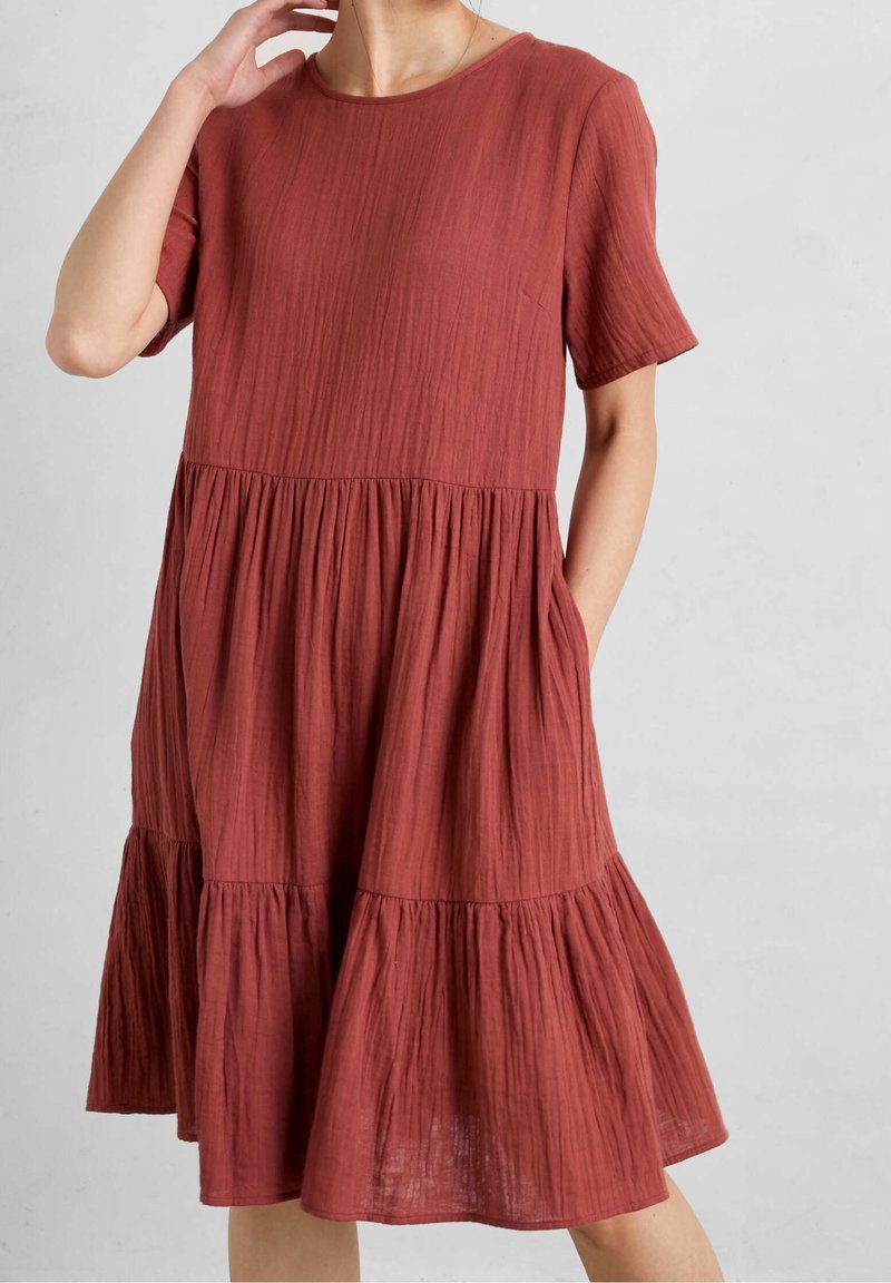Femme portant une robe ample à manches courtes de couleur rouille avec des volants et un tissu texturé, une main dans la poche et l'autre près du cou.