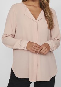 Blouse rose clair avec un tissu doux et fluide. Elle présente un col en V, des manches longues et des poignets boutonnés. L'ourlet est arrondi avec des fentes latérales.