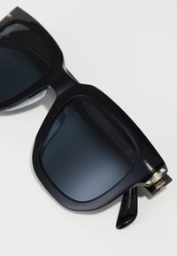 Ochelari de soare supradimensionați, cu lentile întunecate, rame din plastic groase, brățări texturate negre și accente aurii aproape de balamale.