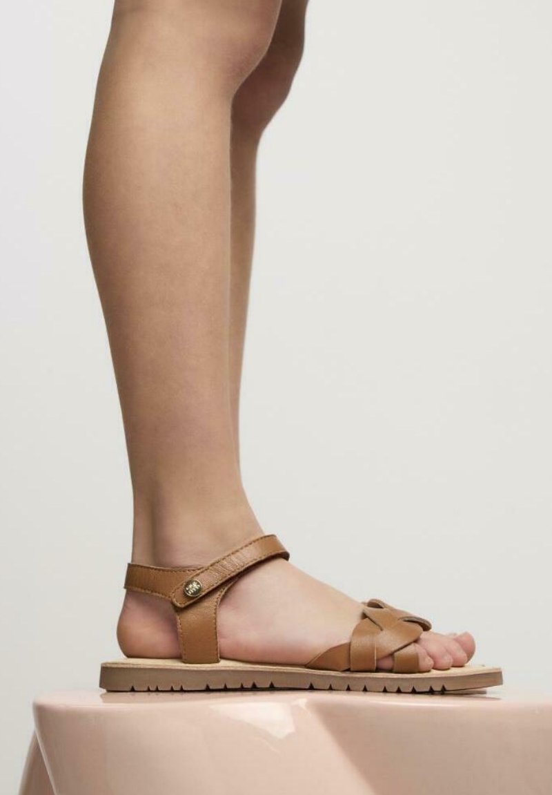 Sandalia de cuero marrón con una correa alrededor del tobillo y diseño entrelazado. Suela plana con fondo texturizado, que muestra los dedos del pie.