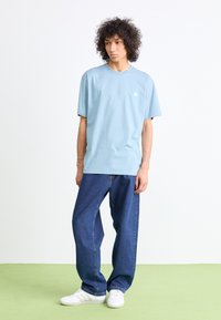 Light blue cotton t-shirt with short sleeves, round neckline, and a small white logo. Paired with loose-fitting dark blue jeans and white sneakers.