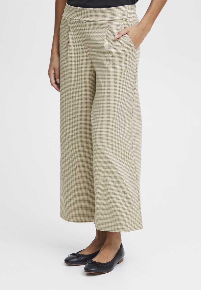 Pantalons larges beiges à motif à carreaux, avec un devant plat, des poches latérales et des détails plissés. Associés à des chaussures plates noires.