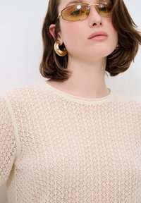 Beiger Strickpullover mit einem detaillierten offenen Muster, rundem Hals und kurzen Ärmeln; getragen mit goldenen Creolen und getönten Sonnenbrillen.