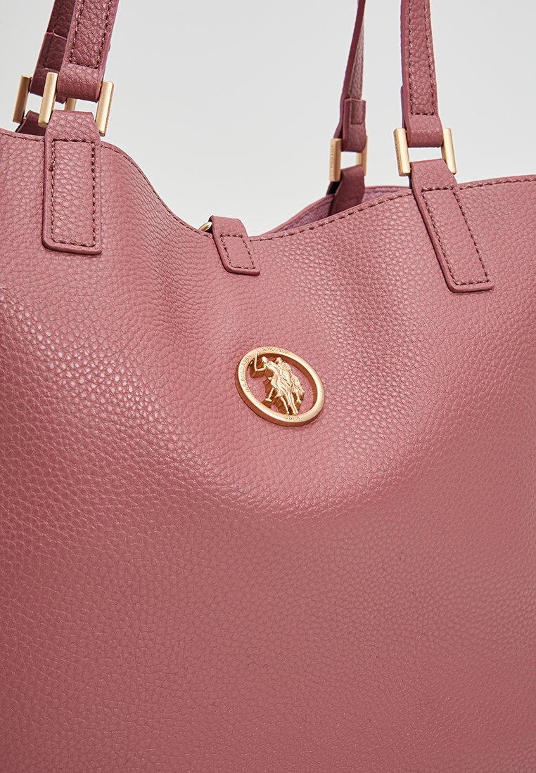 Bolso de mano de cuero texturizado rosa con dos correas, que presenta un acento dorado de logo circular en la parte frontal y una forma suave y redondeada.