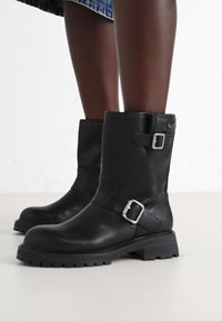 Botas de tornozelo em couro preto com biqueira arredondada, detalhes em fivela e sola com rasto grossa. Apresentam uma textura lisa e um design estruturado.