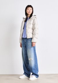 Doudoune gris clair avec capuche, présentant un motif matelassé. Portée sur une chemise bleue et assortie avec un jean large déchiré et des baskets blanches.