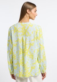 Langärmlige Bluse aus hellblauem Stoff mit gelbem abstraktem Muster. Sie hat einen runden Ausschnitt und eine lockere Passform mit einer Rückenfalte.