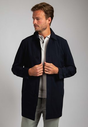 Marinemantel gemaakt van gladde stof, voorzien van een knoopsluiting, lange mouwen en een klassieke kraag, gedragen over een lichte trui.