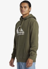 Olive green hoodie made of smooth fabric with a kangaroo pocket, drawstring hood, and a white logo on the front. Classic fit.