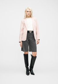 Γυναίκα που φοράει ανοιχτό ροζ σακάκι, λευκή μπλούζα, μαύρη ζώνη με αγκράφα σε σχήμα καρδιάς, γκρι σορτς που φτάνουν μέχρι το γόνατο και μαύρες μπότες μέχρι το γόνατο, όρθια.