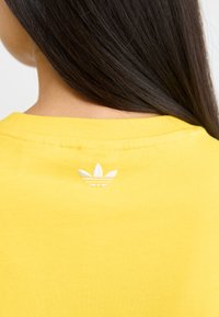 Yellow cotton t-shirt with a ribbed crew neckline and a white embroidered logo on the back. Smooth texture, casual fit.