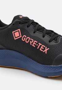 Chaussure de sport noire avec un dessus en tissu texturé, logo GORE-TEX en rose et une semelle en caoutchouc bleue avec un motif de traction.
