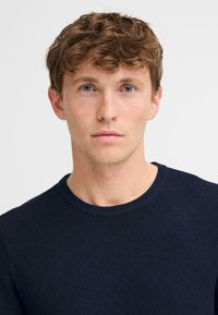 Donker marineblauwe crew-neck trui met een geribde halslijn, een gladde textuur en een aansluitend ontwerp. Model heeft kort, golvend haar en een lichte huidtoon.