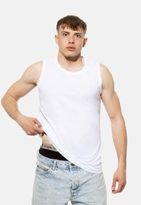 Débardeur sans manches en coton blanc avec un col rond, associé à un jean en denim bleu délavé. Ceinture noire visible avec détail de logo.